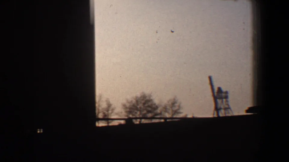 1960 New York City home-movie frame looking from a dark room toward sunset, with bare trees and a small tower-like silhouette outside.