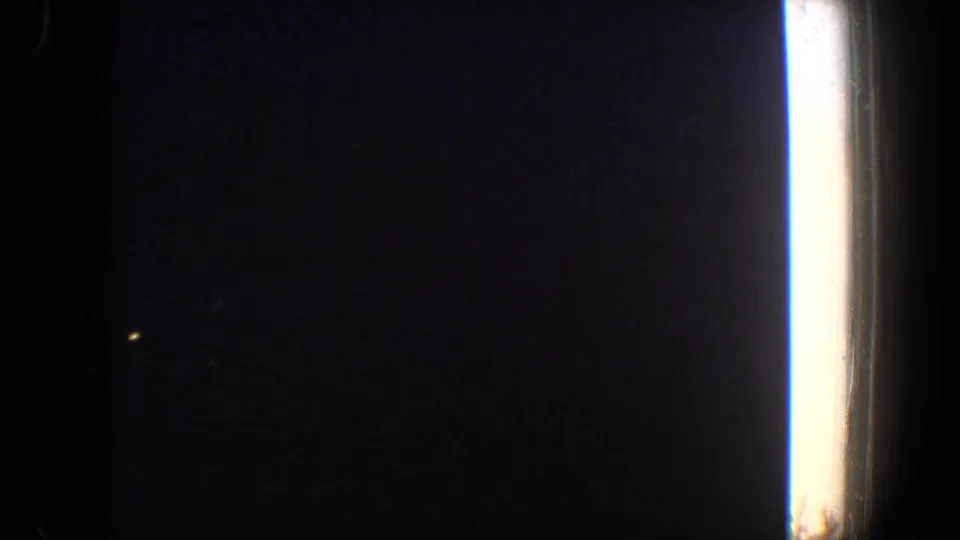 A nearly black home-movie frame from inside a room, showing a bright vertical window edge and a small distant light at dusk.