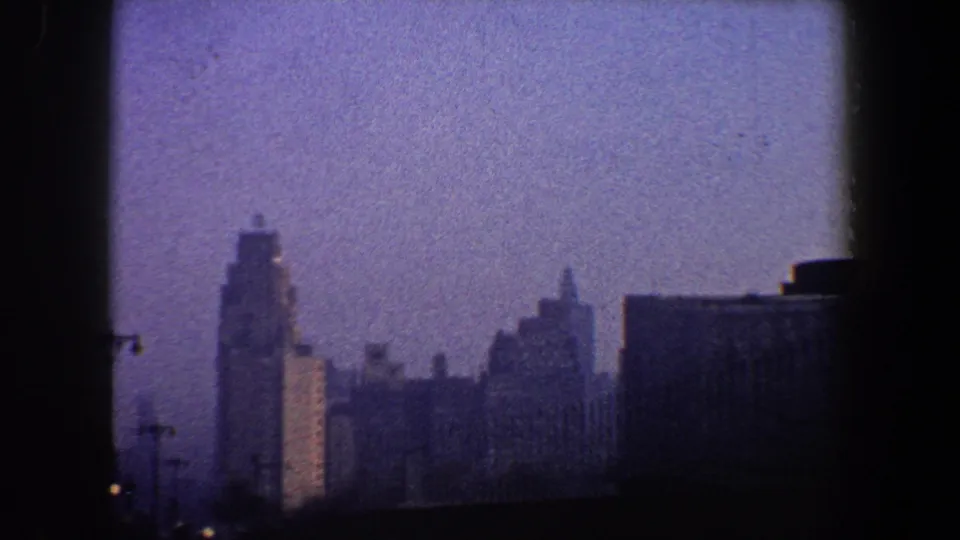 1960 New York City home-movie frame with tall distant buildings, a low rooftop silhouette, and purple-blue haze across the skyline.
