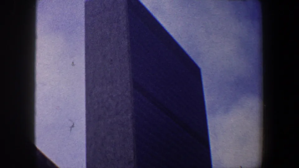 A dark New York City skyscraper fills the frame, its upper facade silhouetted against blue sky in 8mm footage.
