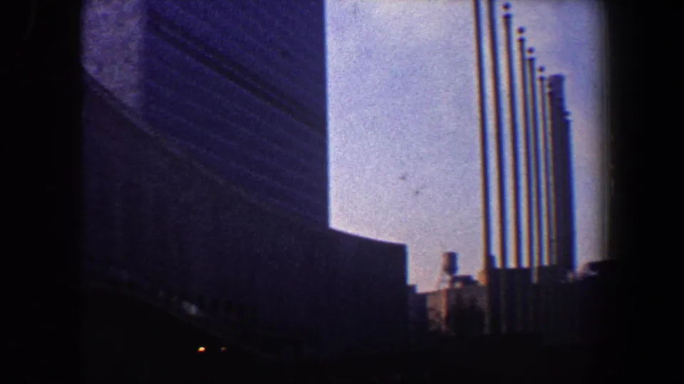 A lower view of the New York City skyscraper shows a row of flagpoles near a curved base, captured on grainy 8mm film.