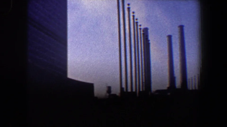 1960 New York City 8mm frame showing a modernist building edge, clustered flagpoles, and a distant smokestack in deep shadow.