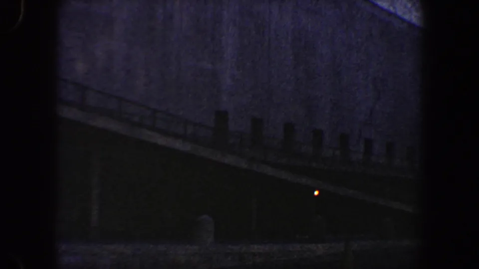 Shadowed New York City exterior with a sloped ramp or walkway, railing posts, large wall surface, and a small red light.