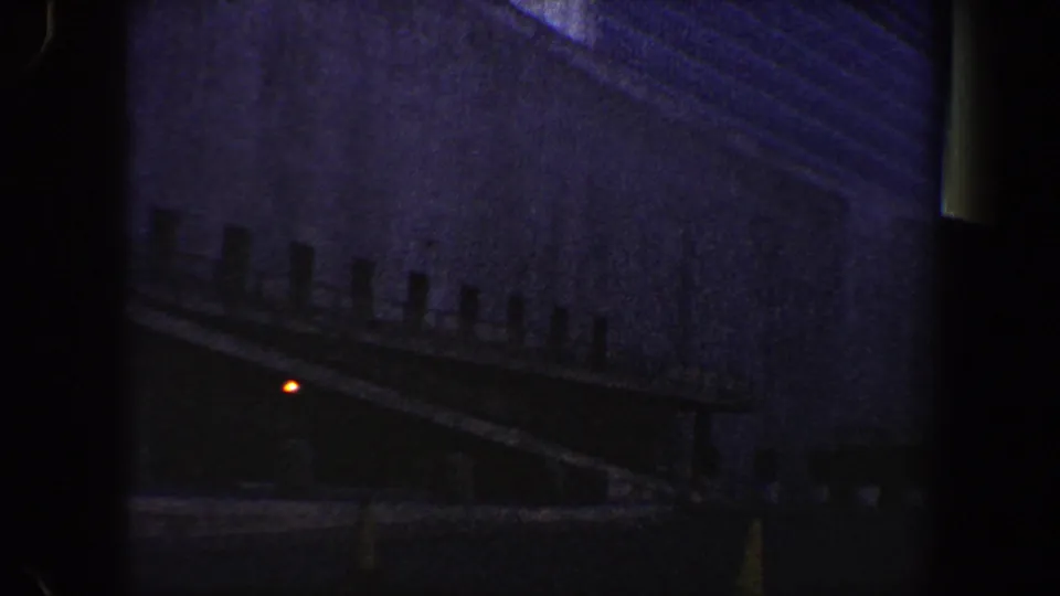 Grainy 8mm view of a dark exterior ramp or walkway, railing posts, building wall, and single glowing red light.