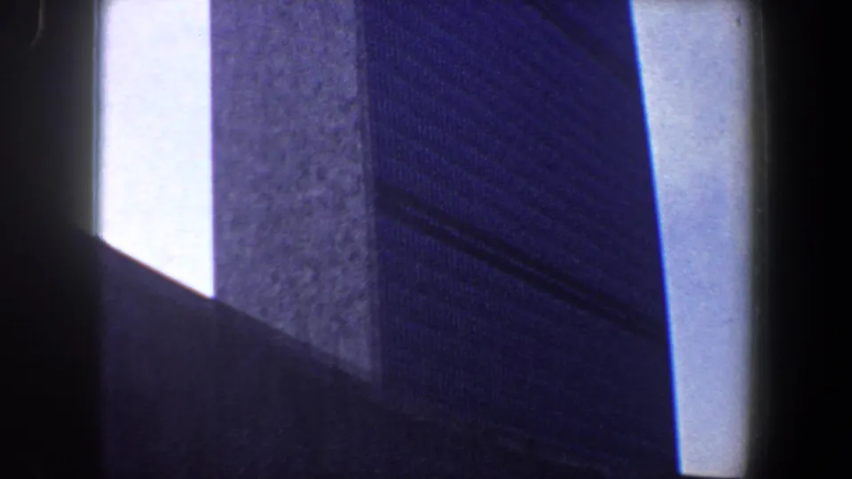 1960 New York City 8mm frame with a textured high-rise wall, deep shadow, and pale sky visible along the building edge.