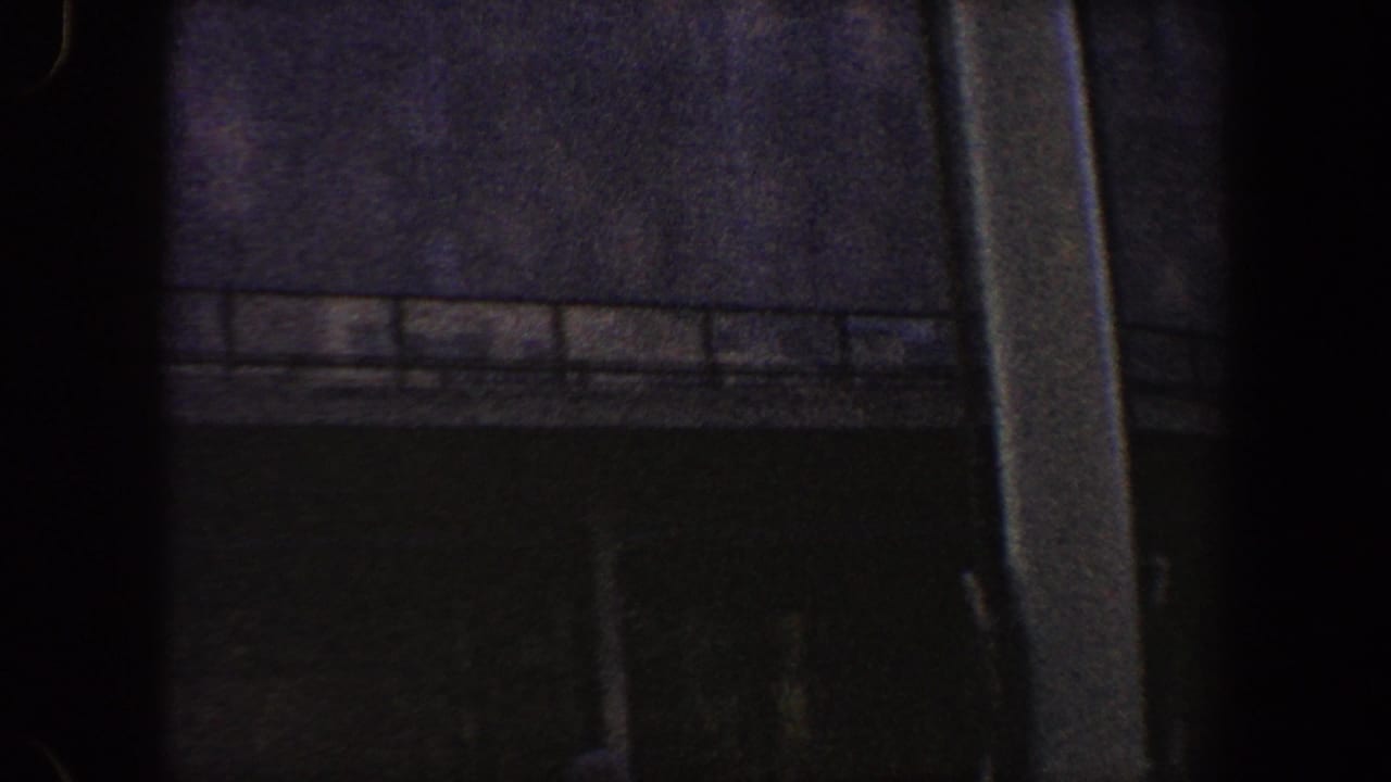Underexposed 1960 New York City 8mm frame showing a dark commercial facade, railing, and heavy vertical foreground shadow.