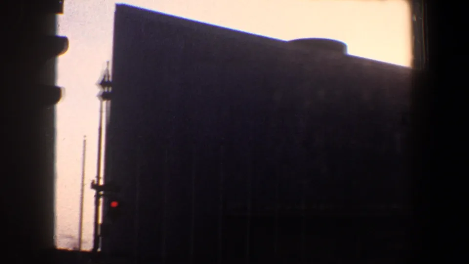 Grainy 1960 New York City frame showing a large dark building facade, red traffic signal, and narrow vertical street fixture.