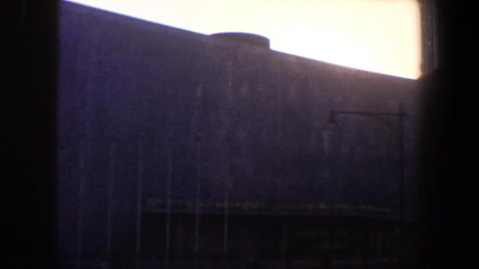 1960 New York City home-movie frame showing a large modernist-looking building with a curved roofline, partly obscured by dark window mullions and bright sky.