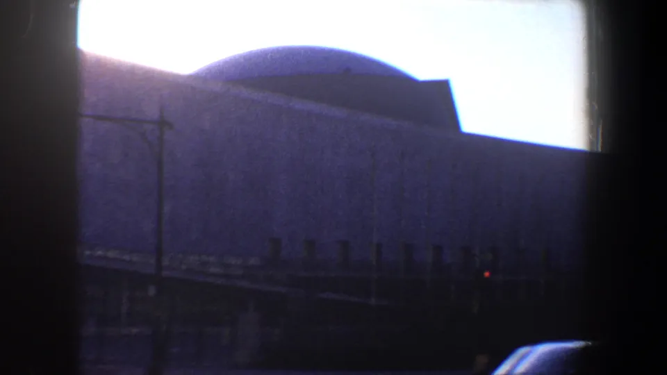 1960 New York City home-movie frame showing the building's curved roof and wall surface, a streetlamp, dark foreground framing, and a small red traffic light.