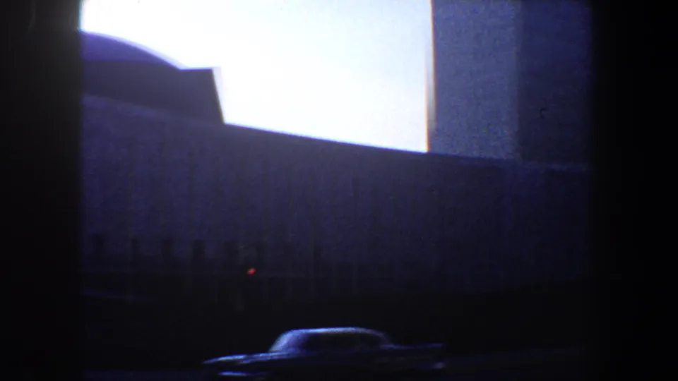 Final 1960 New York City frame with the large building behind dark foreground framing and a car passing at the lower edge of the street view.