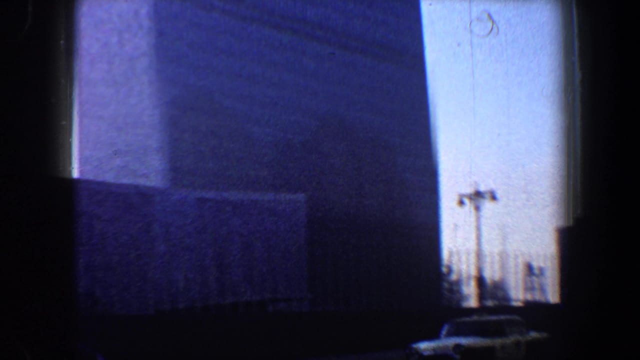 Tall building wall seen from below with sky, road edge, poles, and a small car at lower right.