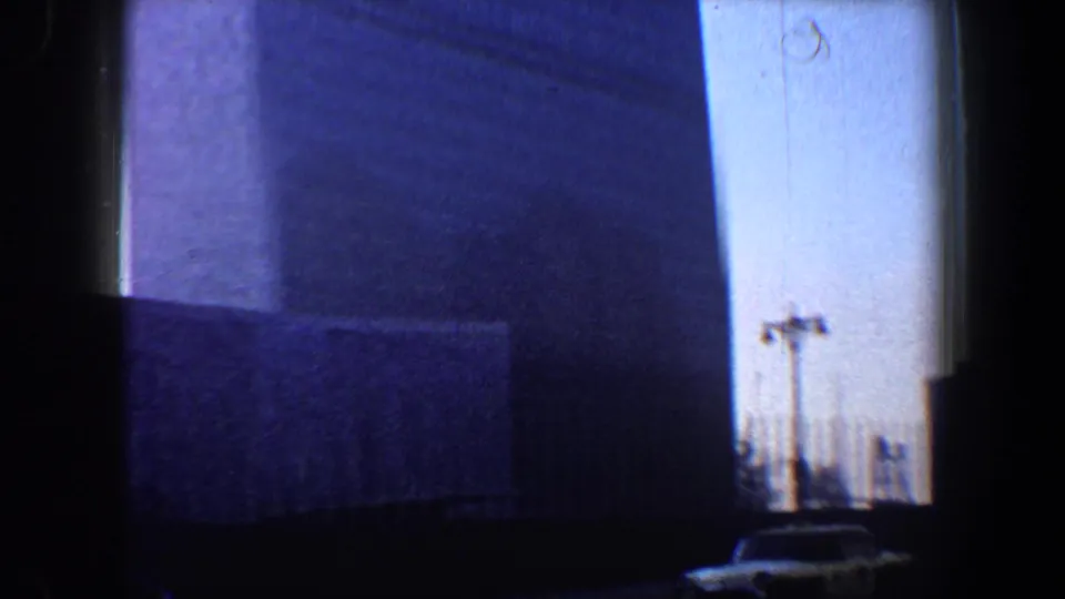 Low-angle 1960 New York City street view of a high building facade, with a road edge, poles, and a car near the bottom of frame.