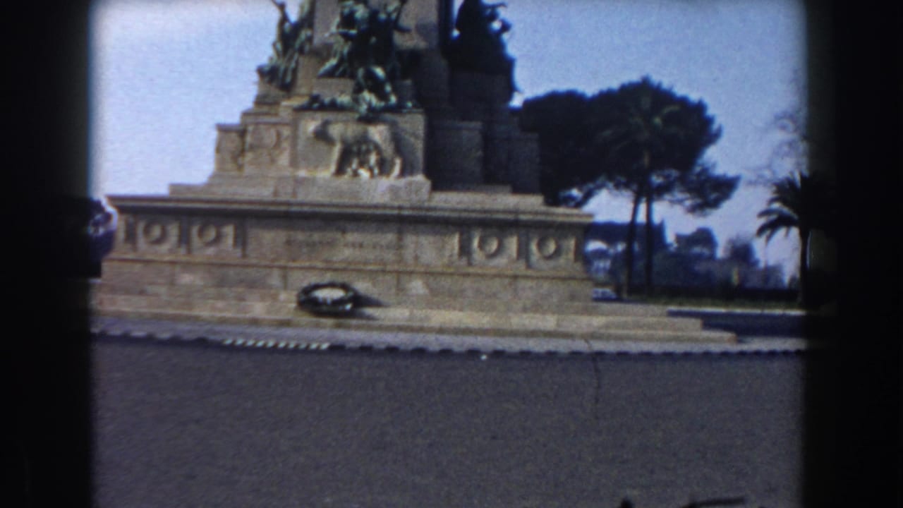Stone monument base with bronze figures, wreath, trees, and sky seen across a road.