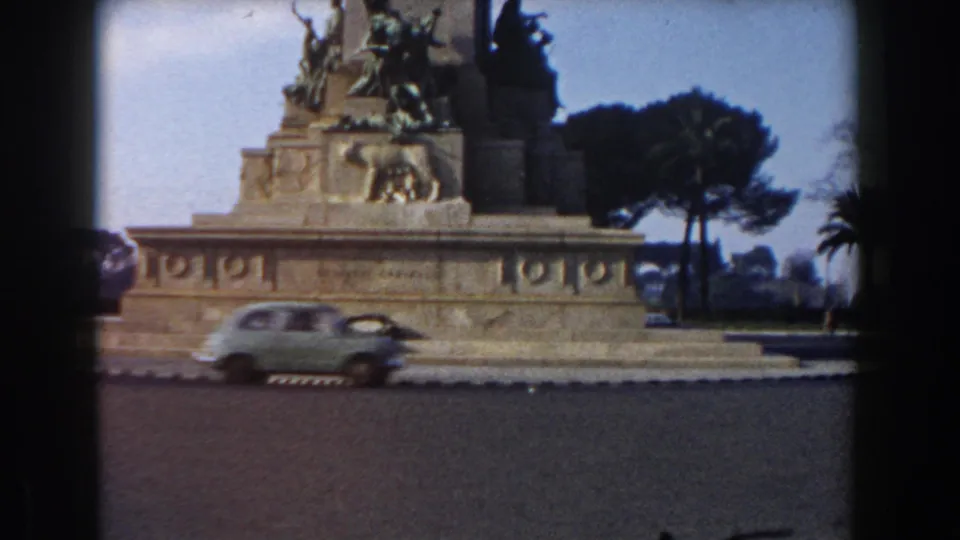 A 1959 Rome travel frame catches a small car crossing the road in front of the stone monument and bronze figures.