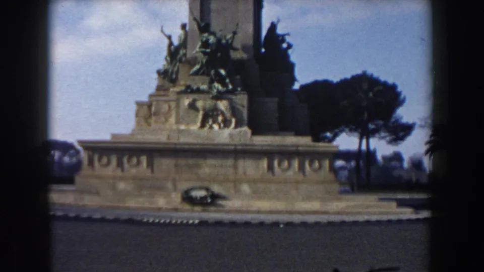 Final Rome 1959 8mm frame holding on the monument, with bronze figures, a wreath, road foreground, and trees in soft color.