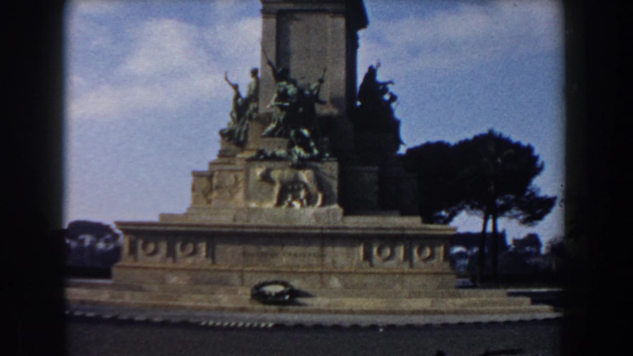Stone monument base with bronze sculptures, lion relief, wreath, trees, and blue sky.