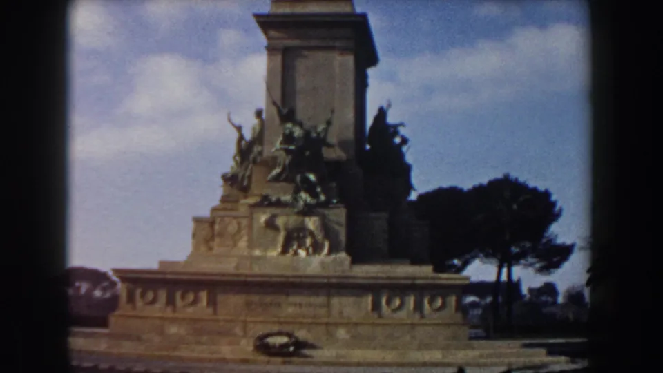 Rome travel-film frame with a stone memorial base, bronze sculptural groups, a lion relief, a wreath, and a dark tree silhouette.