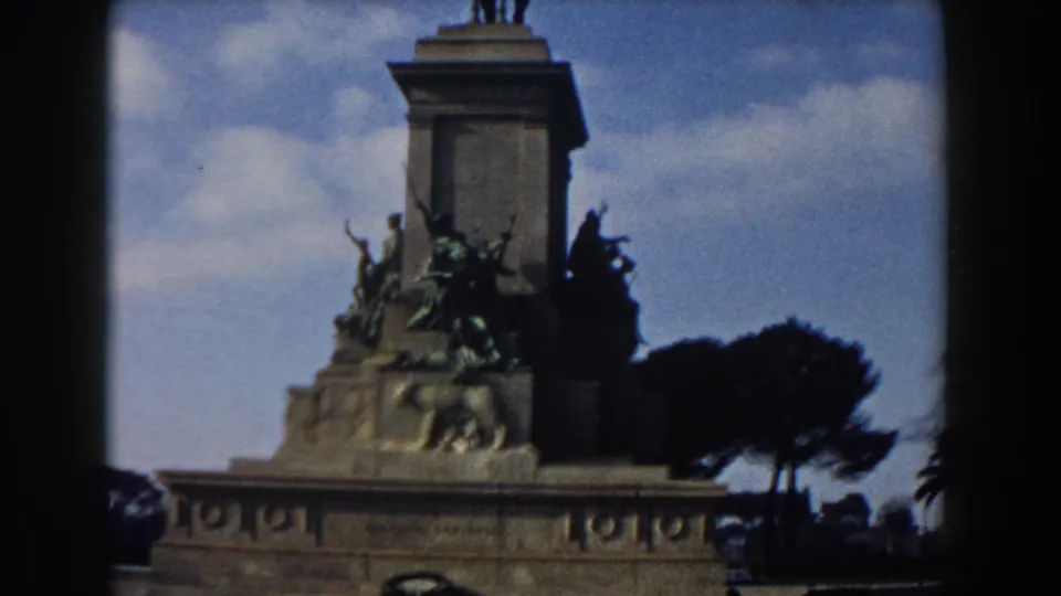 1959 Rome frame during an upward tilt, showing bronze figures on a stone pedestal beneath a tall column and partly cloudy sky.