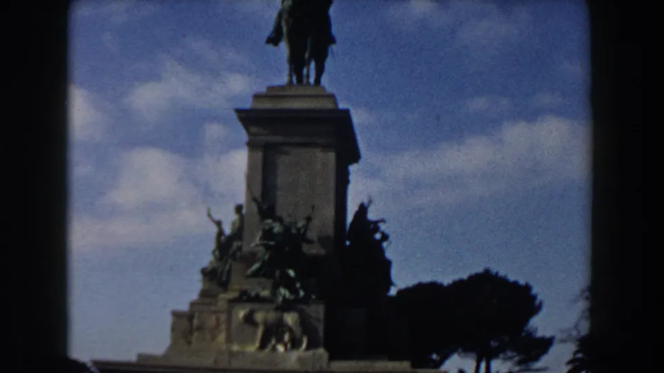 Rome 8mm frame showing more of the tall monument column, with a statue on top, bronze figures below, trees, and blue sky.