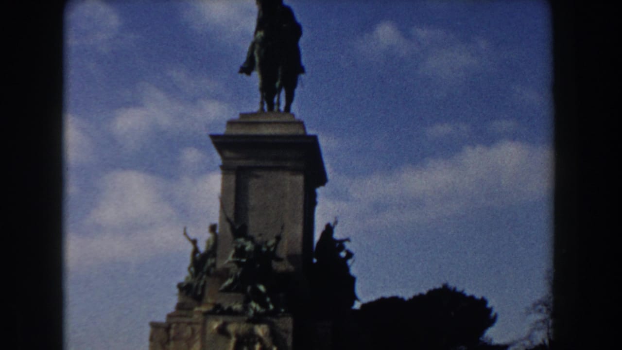 1959 Rome home-movie frame showing a dark equestrian monument above a carved stone pedestal, partly framed by 8mm film borders.