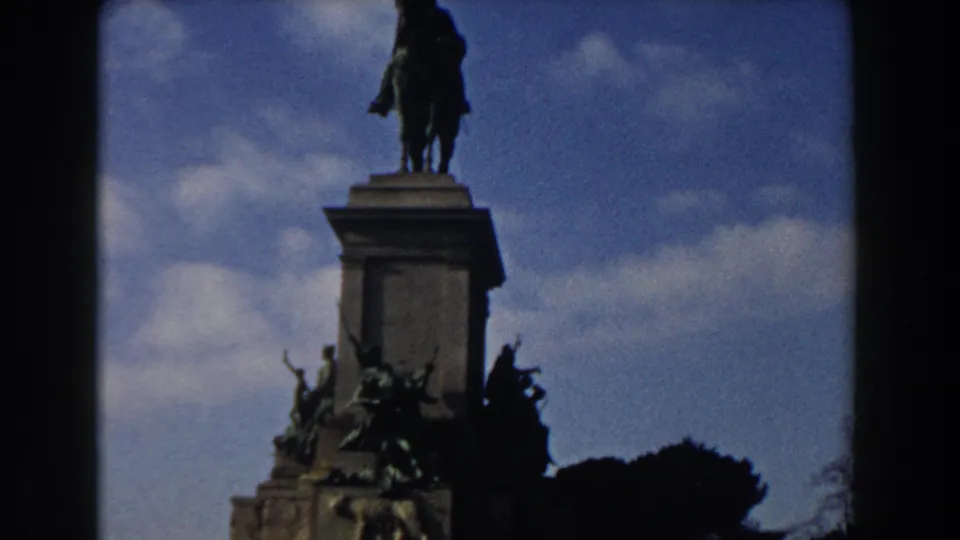 1959 Rome home-movie frame showing a dark equestrian monument above a carved stone pedestal, partly framed by 8mm film borders.