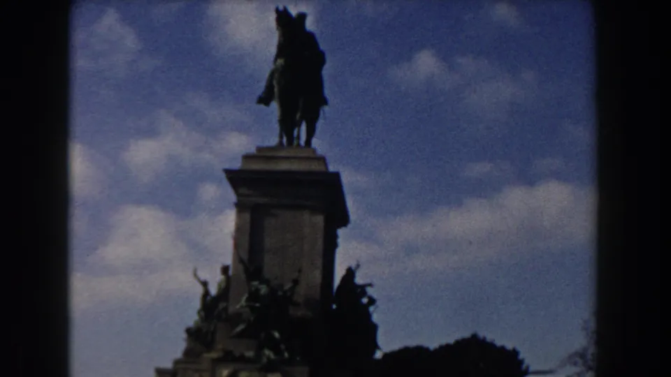 1959 Rome frame showing the monument from below, with pedestal carvings, tree silhouettes, and clouds surrounding the statue.