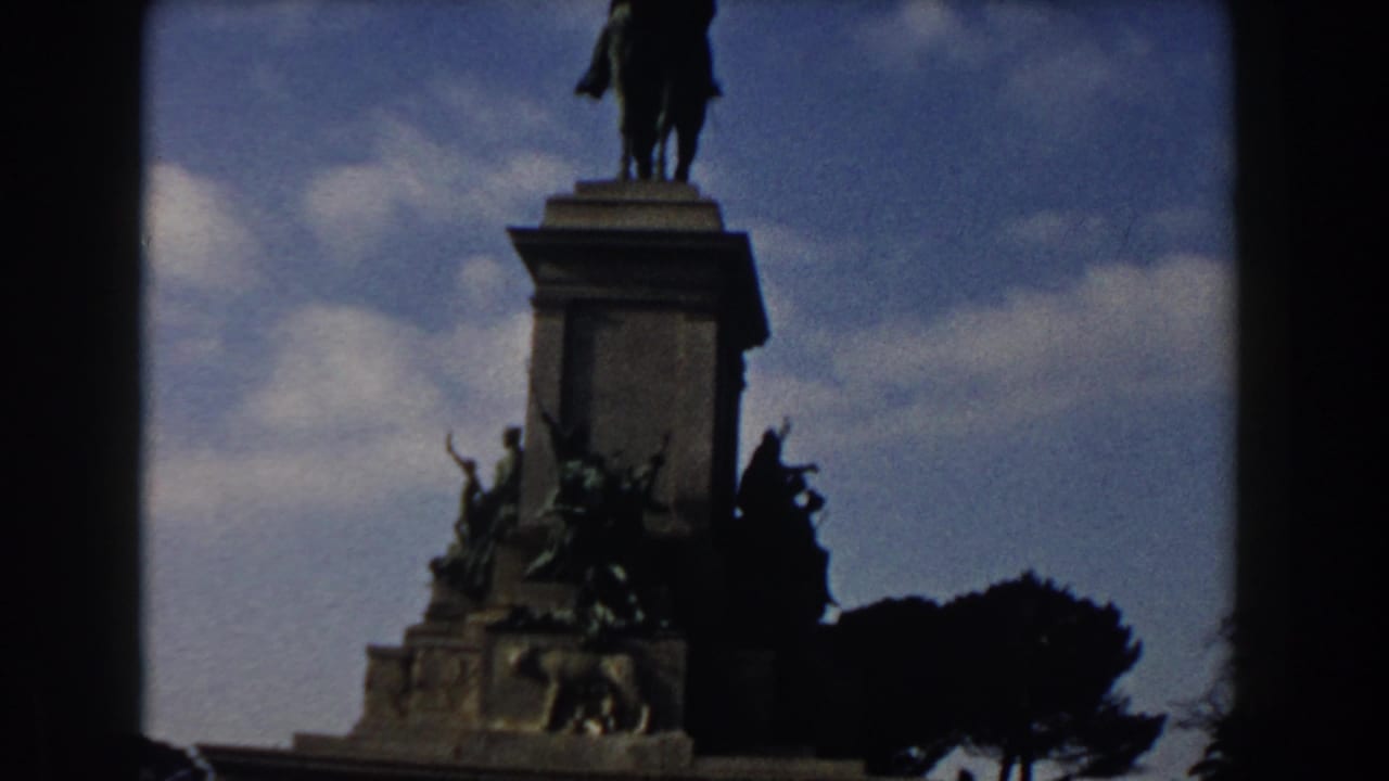 1959 Rome home-movie frame showing an equestrian monument, sculpted base, umbrella pines, and blue sky.