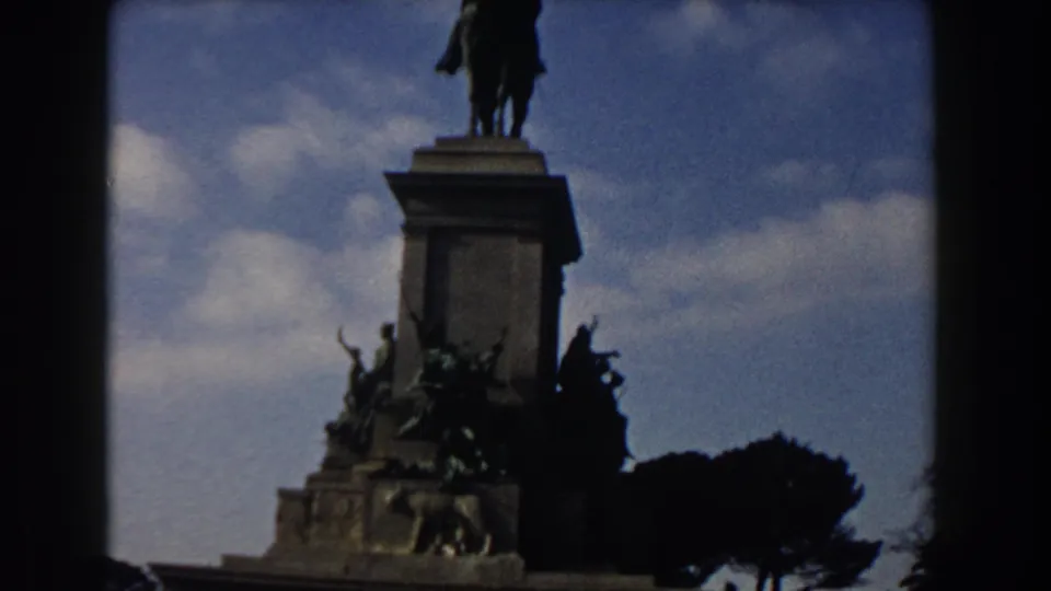 1959 Rome home-movie frame showing an equestrian monument, sculpted base, umbrella pines, and blue sky.