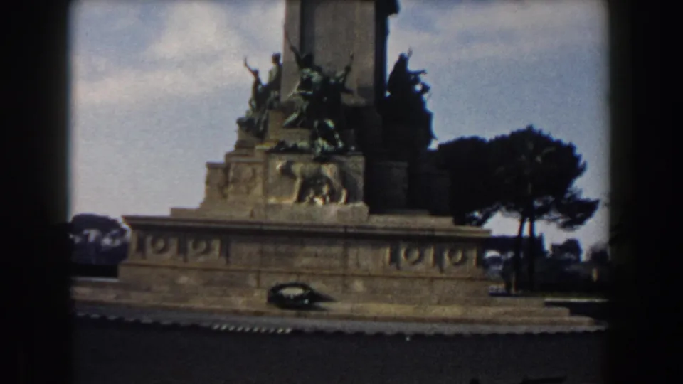 1959 Rome frame showing a decorated equestrian monument, street edge, trees, and a dark classic car passing below.
