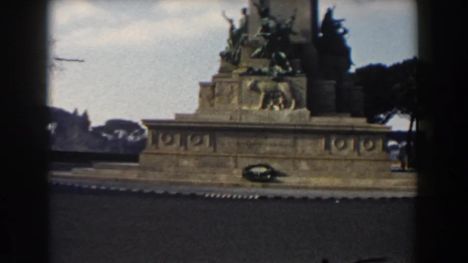 Rome home-movie frame with a dark car crossing the foreground beneath a sculpted equestrian monument and umbrella pines.