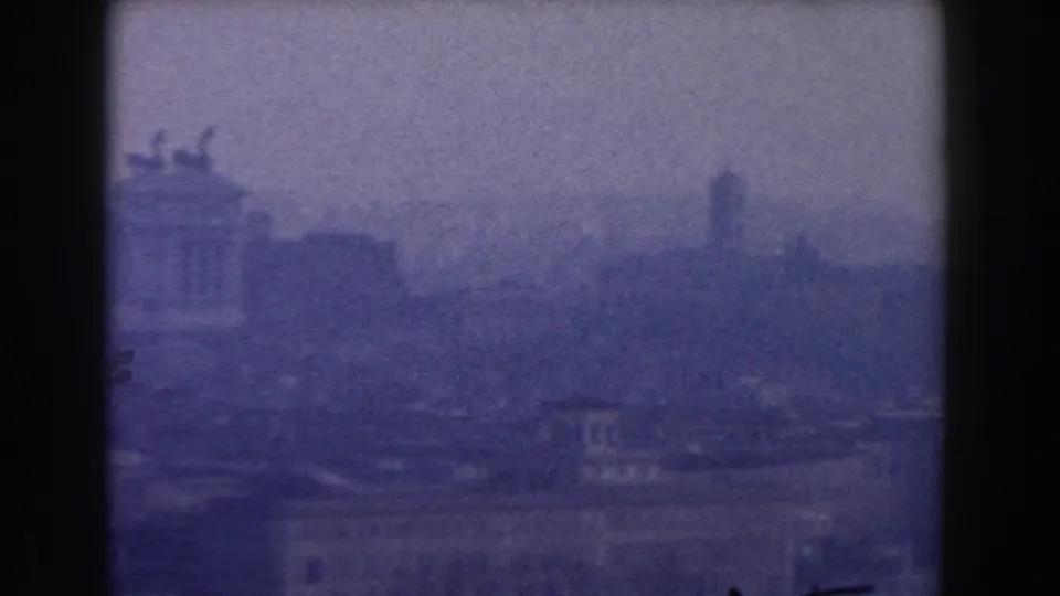 Elevated 1959 Rome view with rooftops in the foreground and a tall distant form softened by haze.