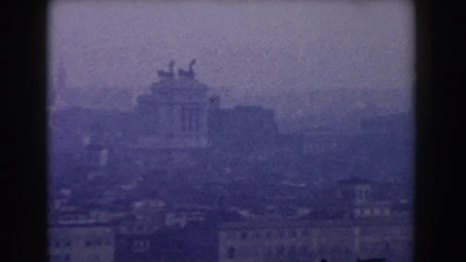 1959 8mm view of Rome through haze, with pale buildings and a classical rooftop silhouette faintly visible.