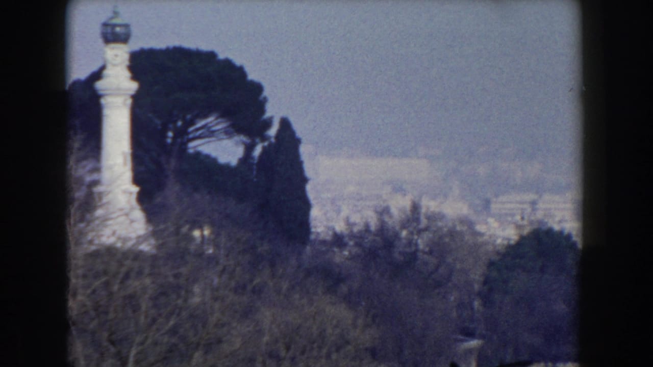 1959 Rome frame with a pale monument or column beside umbrella pines, set against a hazy distant city backdrop.