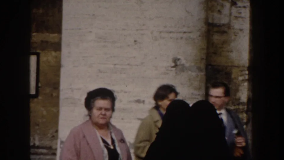 Rome travel footage shows tourists, including a woman in a pink coat and a man in a suit, near monumental stonework.