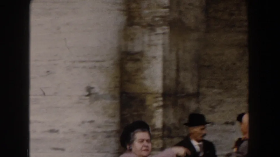 A late-1950s Rome visitor scene shows tourists moving past weathered stonework, with a man in a dark hat at the edge of the frame.