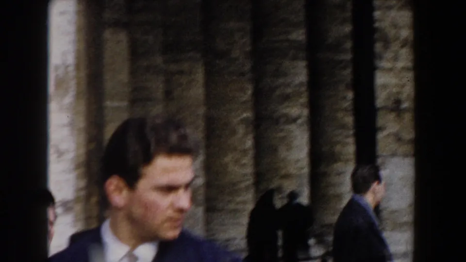 Close 1959 Rome frame of a suited man near a church exterior, with other adults moving among tall stone columns.