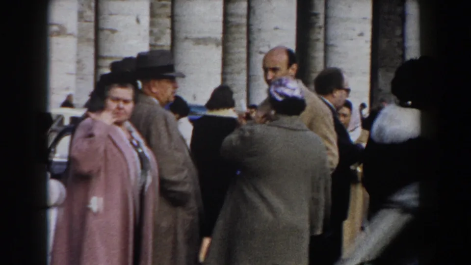 Rome 1959 frame showing a clustered public crowd in coats before monumental columns, with a woman in pink at left.