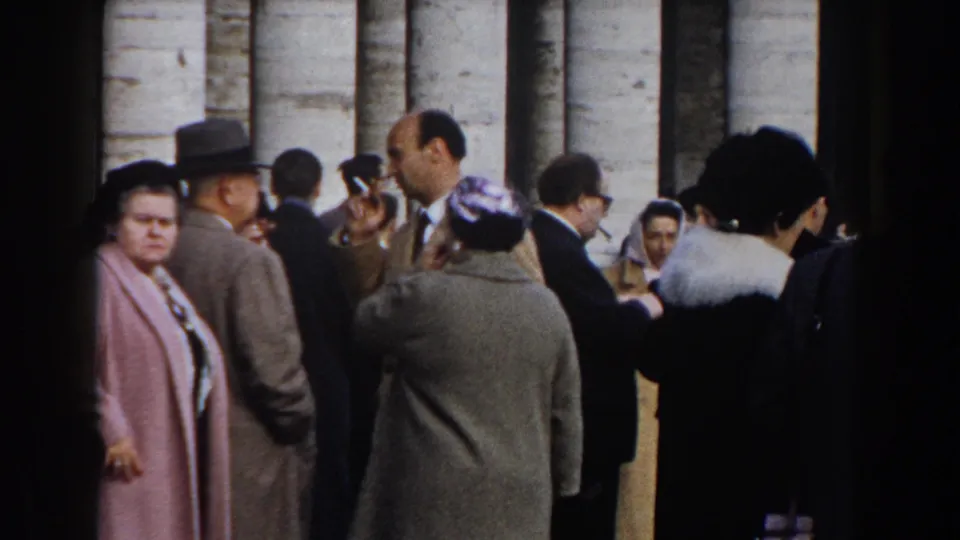 Rome 1959 home-movie frame of visitors near a colonnade, including adults in hats and coats and a man raising a camera.
