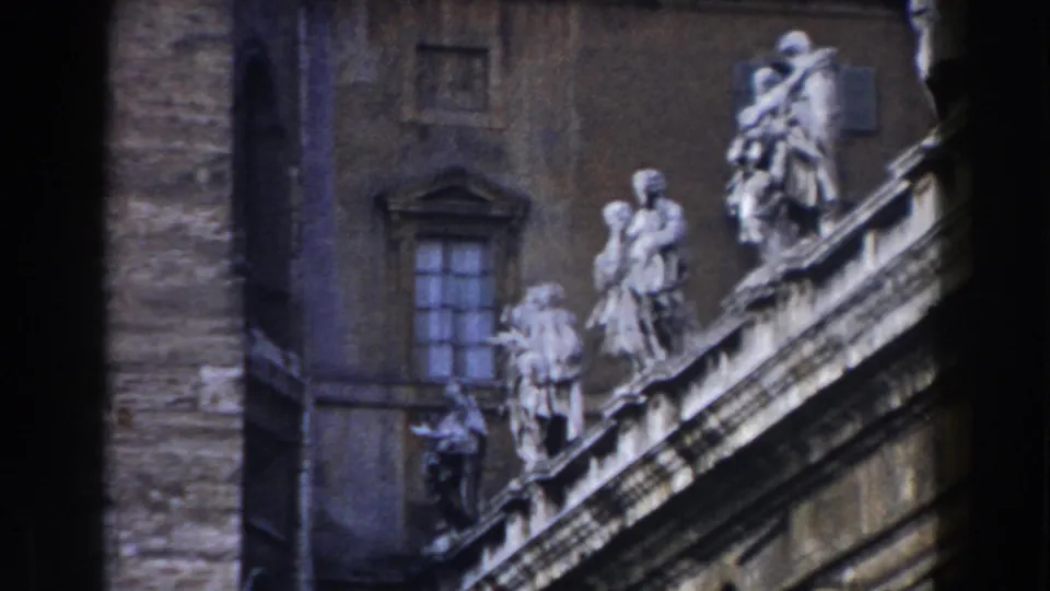 1959 Rome travel footage frame with classical stone figures along a roofline and a tall facade behind them.