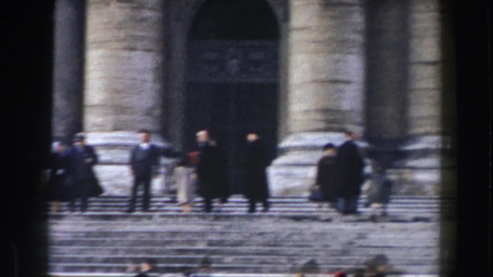 Visitors in mid-century attire gather on steps outside a monumental stone entrance in Rome, Italy.
