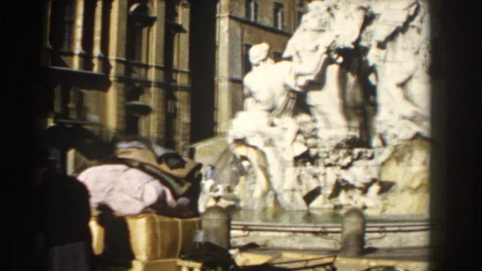Sunlit Roman fountain scene with classical sculpture, street architecture, bollards, and a nearby pedestrian.