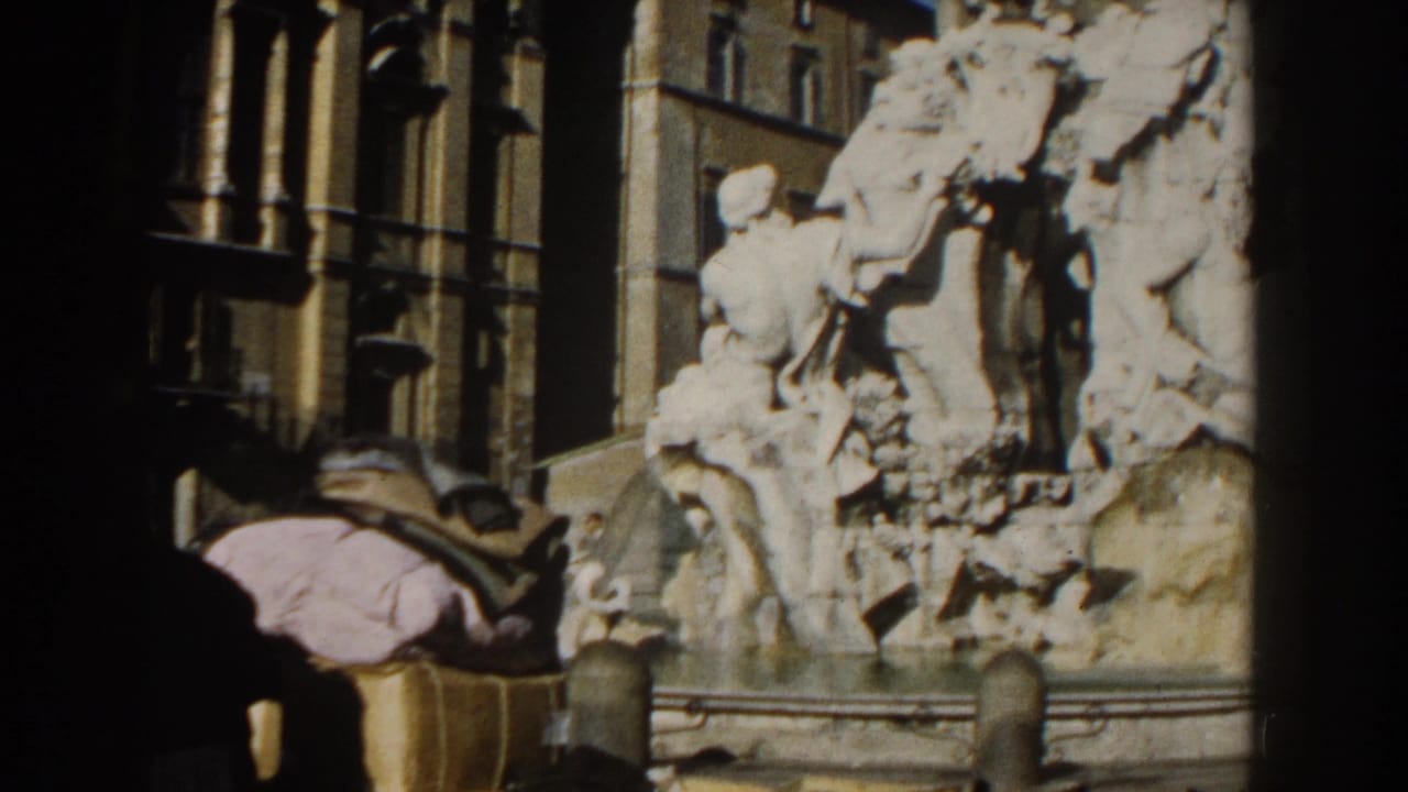 Stone fountain sculpture beside a Roman building with part of a parked vehicle at the edge.