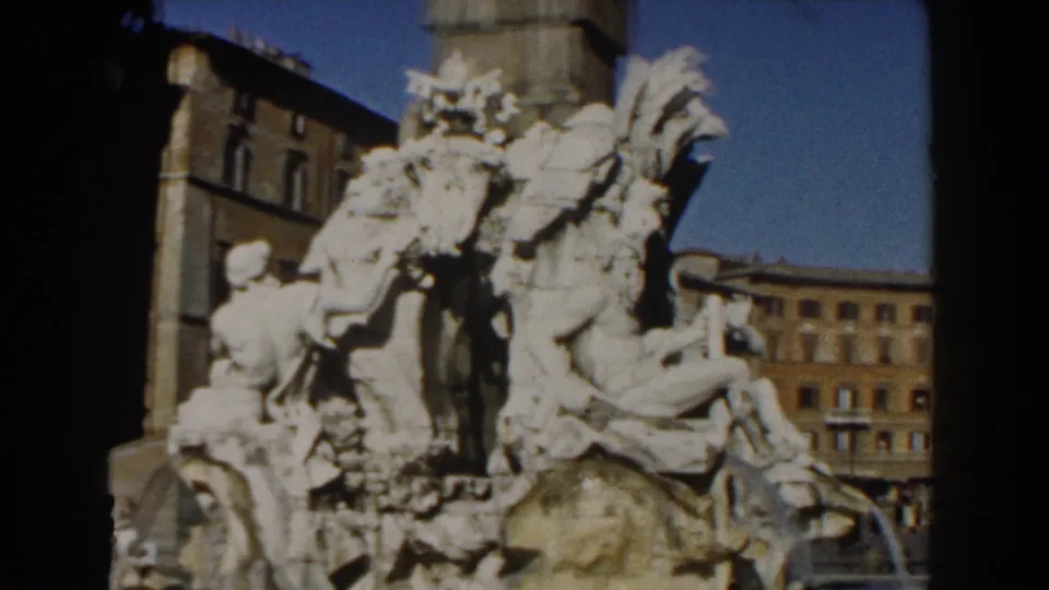 1959 8mm Rome view of a monumental fountain with carved figures set against urban facades and clear blue sky.