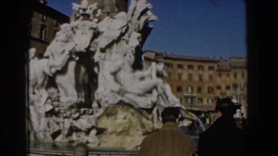 1959 8mm Rome frame with pedestrians in mid-century coats passing a monumental stone fountain in a public square.