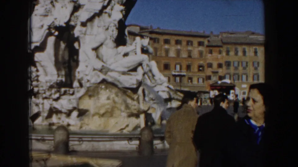 1959 8mm Rome view of visitors near the stone fountain, with a woman at the right edge and buildings across the square.
