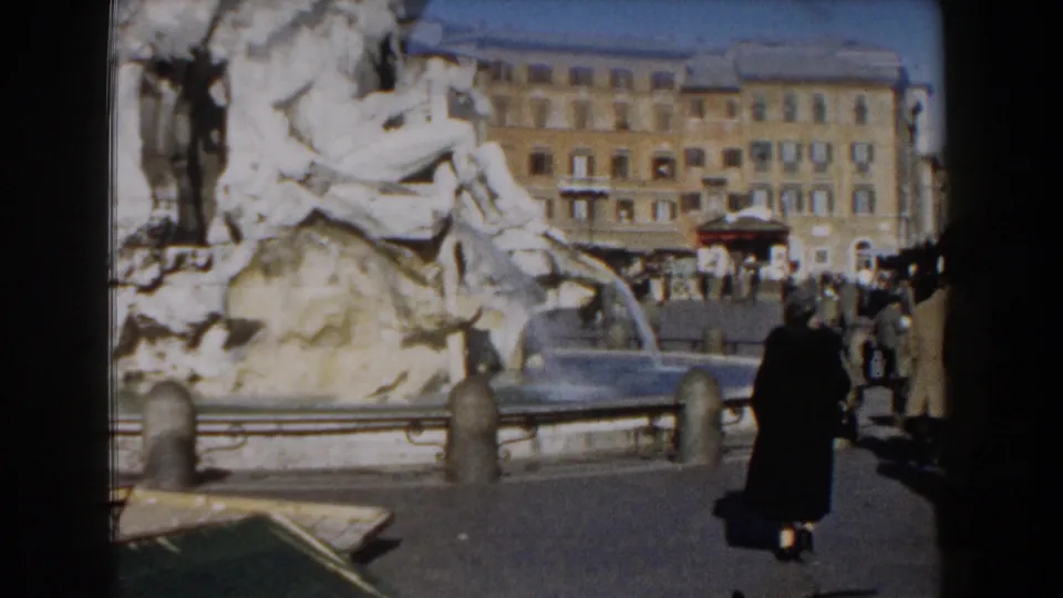 1959 8mm view of the Trevi Fountain in Rome with stone figures, flowing water, pedestrians, and buildings around the piazza.