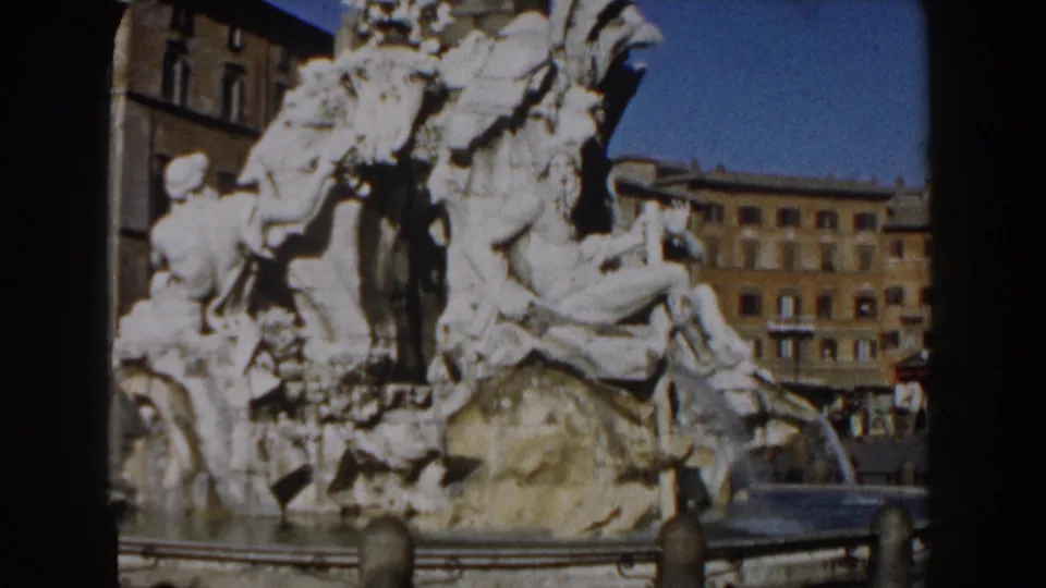 An angled 1959 8mm frame shows Trevi Fountain sculptures, water, foreground bollards, and sunlit buildings beyond the piazza.