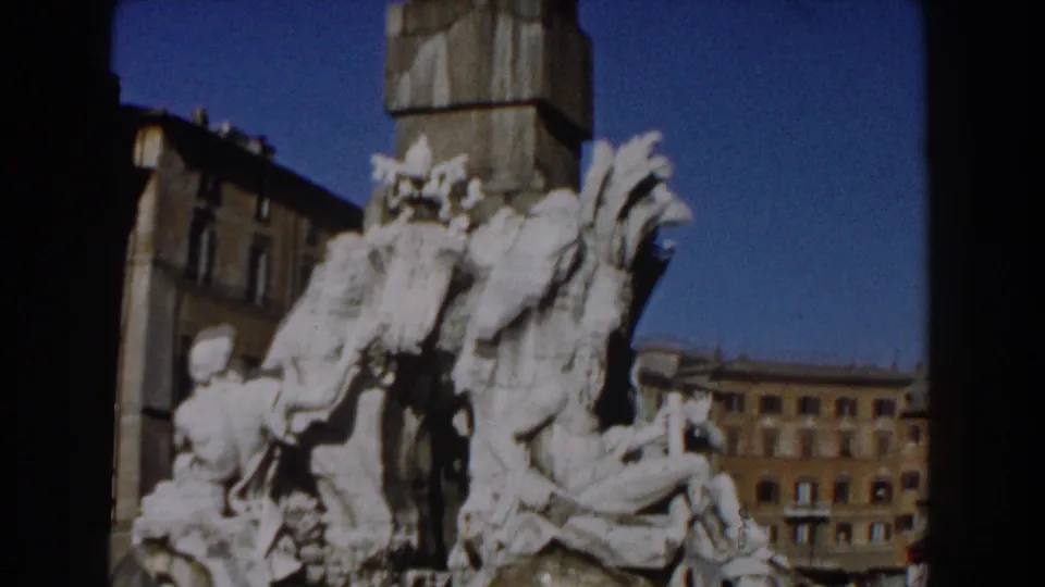 1959 Rome home-movie frame shows the Trevi Fountain sculptures from a tighter angle, with blue sky and building facades nearby.