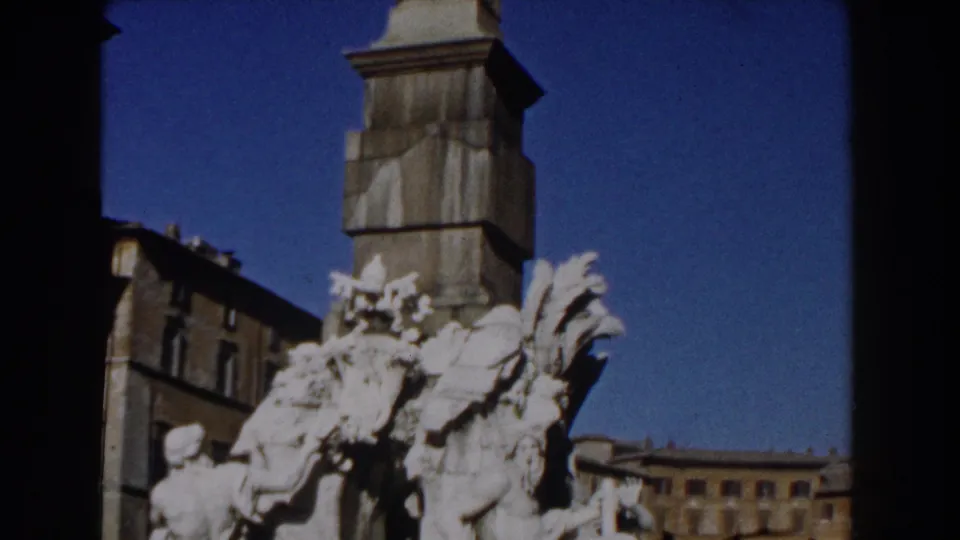 1959 Rome 8mm frame showing ornate pale sculpture near the base of a tall column, with city buildings and blue sky behind it.