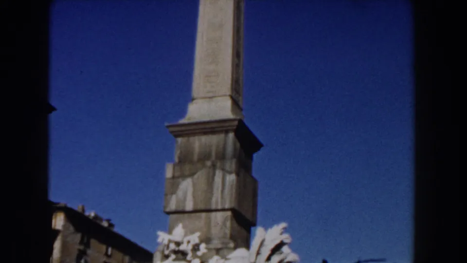 1959 Rome 8mm frame with the column rising through the center, pale sculpture partly visible below, and blue sky in the background.